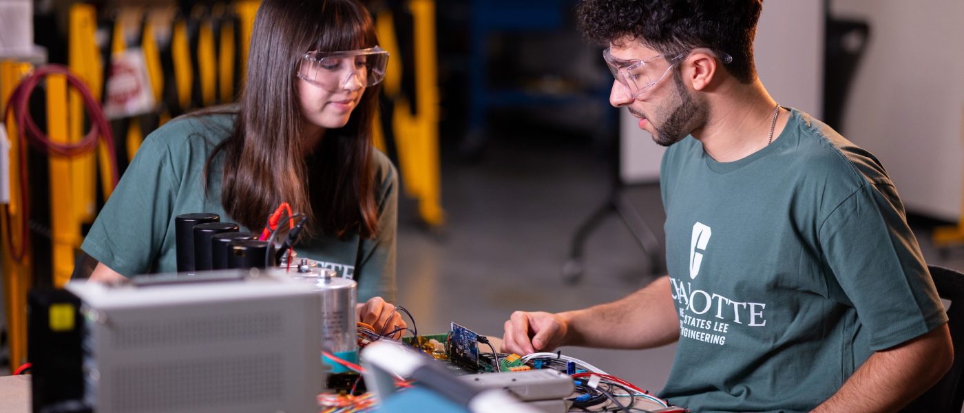 Two students wearing green shirts, working on an electrical project at a table.