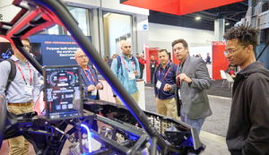 At a trade show, a group of five men are gathered around the open chassis of an electric vehicle. One man in a grey blazer is gesturing toward the vehicle while speaking to the others.