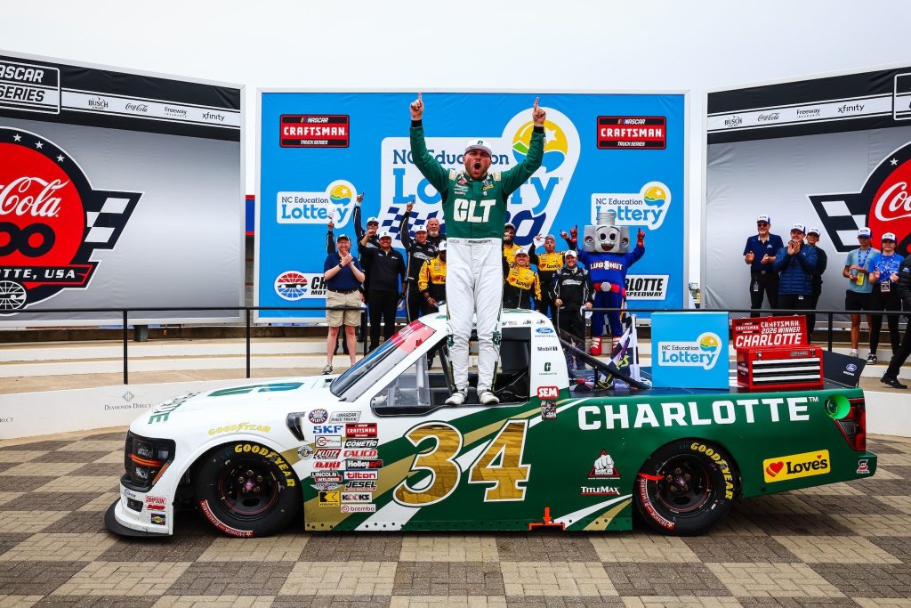 Layne Riggs celebrates NASCAR Craftsman Truck Series win by standing in window of his No. 34 truck. His crew stands behind him.